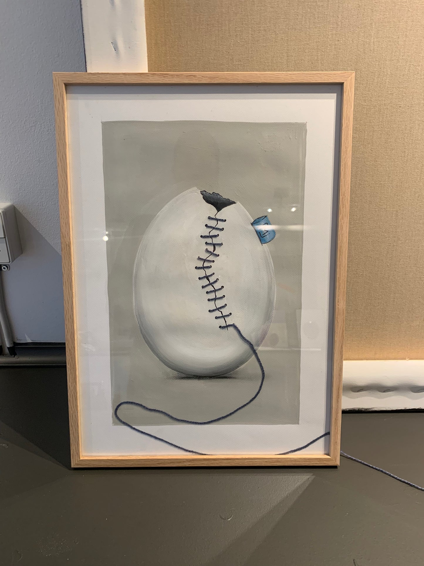 Unikt kunstværk: Hvidt æg med revne, syet sammen med mørkgrå bambusgarn på lysegrå baggrund. Dette originale maleri er udført med akryl, blyant og tusch på 200 g artist quality papir i A3 størrelse. Ægget er hvidt med sort indeni og prydes af et lyseblåt ML tag, hvilket giver en elegant kontrast og tilføjer dybde til kunstværket. Værket leveres uden ramme, så du kan arrangere garnet efter eget ønske og skabe et personligt udtryk. Perfekt til dig, der søger unik, moderne kunst med et råt, men stilrent touch.