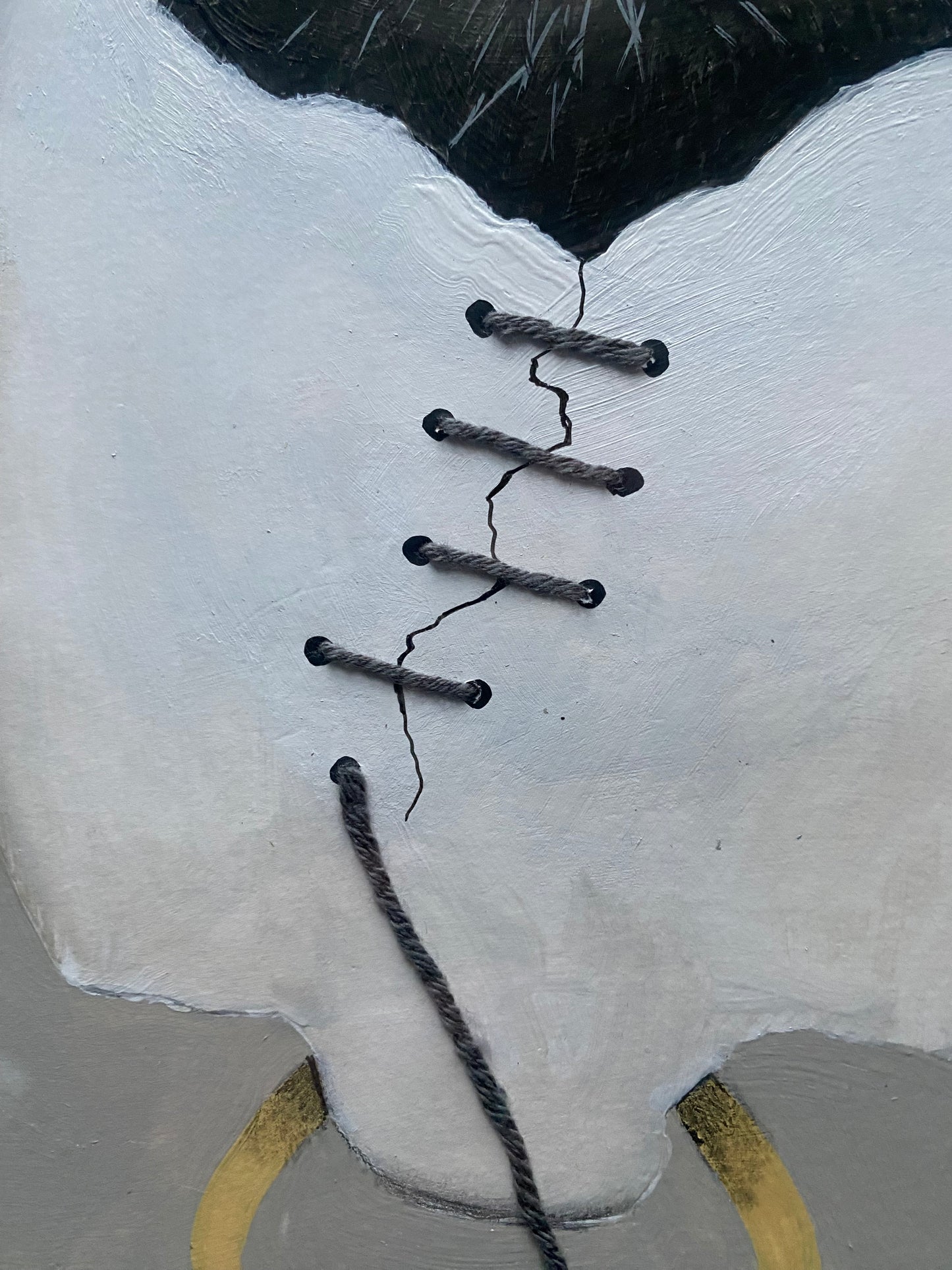 Indrammet kunstværk med et hvidt, revnet æg, der står på gule fødder. Ægget er syet sammen med mørk tråd, og en løs tråd fortsætter ned foran billedet. Øverst ses mørkt, børsteagtigt fyld. Baggrunden er afdæmpet grå.