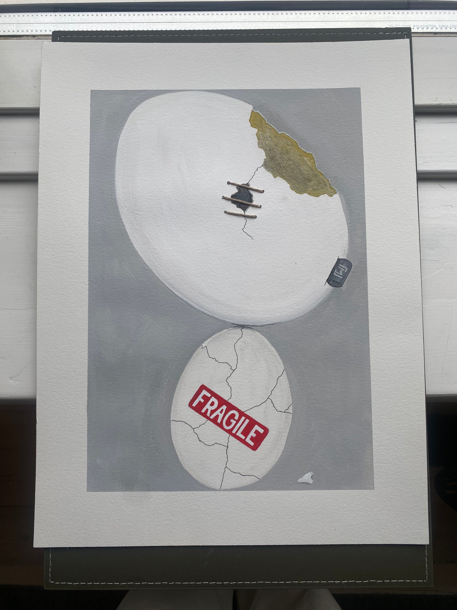 Kunstværk med to æg oven på hinanden. Det nederste æg er revnet og mærket “fragile”. Det øverste æg er større og delvist repareret med synlig tråd og et gyldent brud.