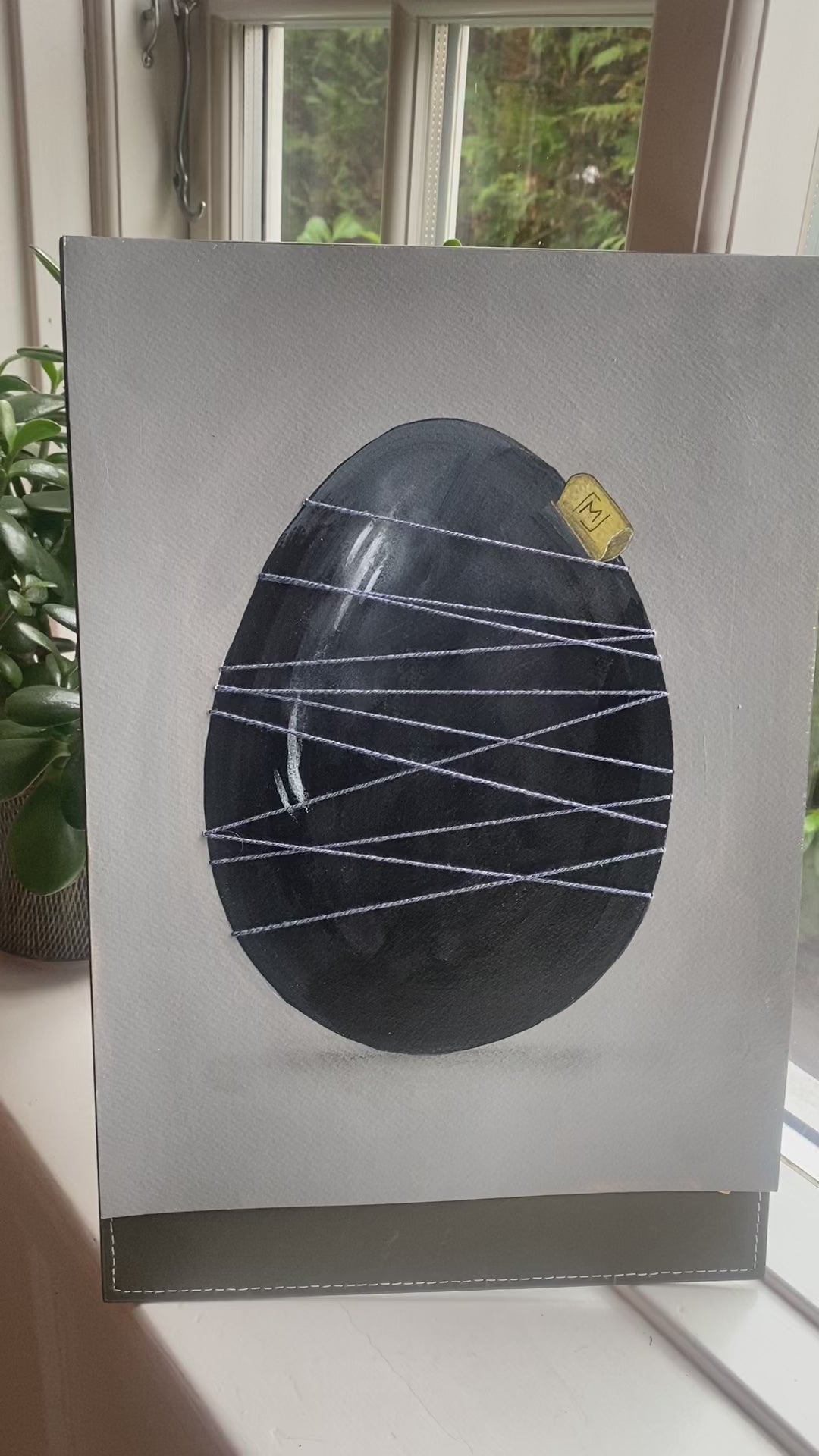 Mørkt håndmalet æg holdt sammen af blå tråd. Original kunst med roligt udtryk, synlige lag og fokus på materialitet og balance.