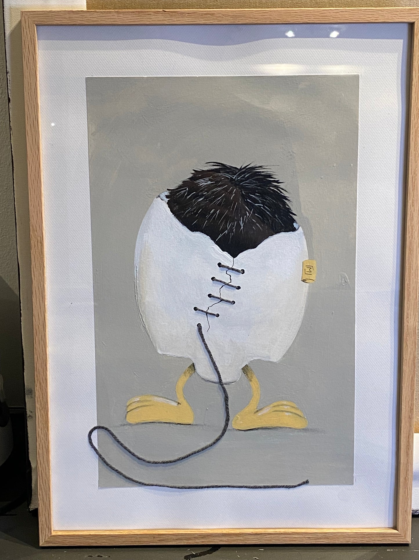 Indrammet kunstværk med et hvidt, revnet æg, der står på gule fødder. Ægget er syet sammen med mørk tråd, og en løs tråd fortsætter ned foran billedet. Øverst ses mørkt, børsteagtigt fyld. Baggrunden er afdæmpet grå.